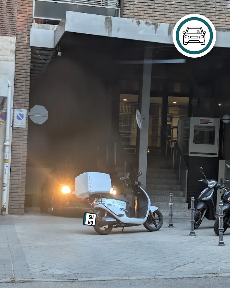Moto aparcada delante de la salida de un garaje con vado; dentro, un coche con los faros encendidos espera para salir